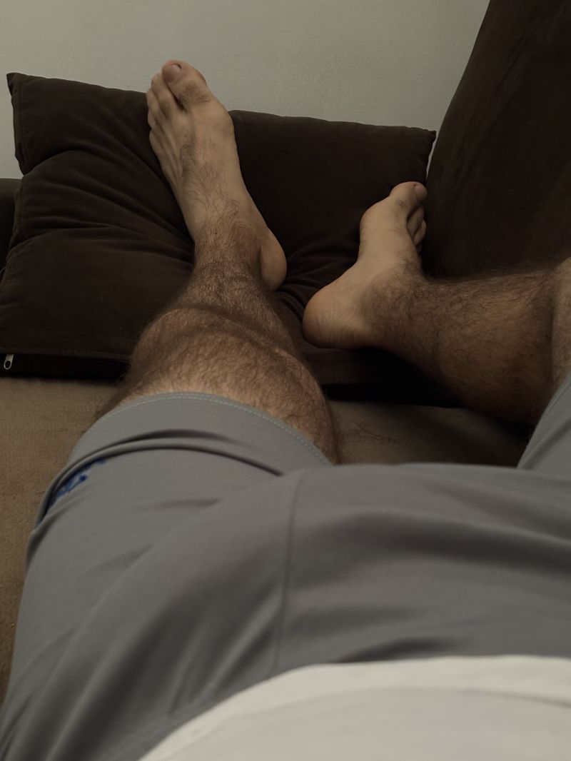 Deitado no sofá mostrando a mala no meio das pernas e o pezão peludo